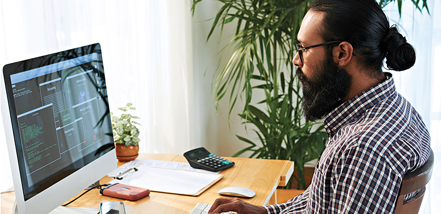 Contemporary programmer in casualwear sitting by desk in front of computer monitor while developing new software