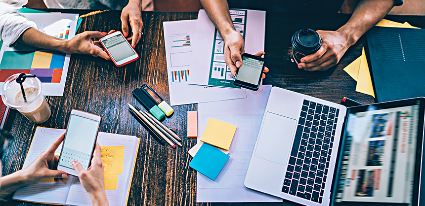 Crop view from above of modern colleagues using mobile phones while sitting with notepads and papers at table in cafe having drinks